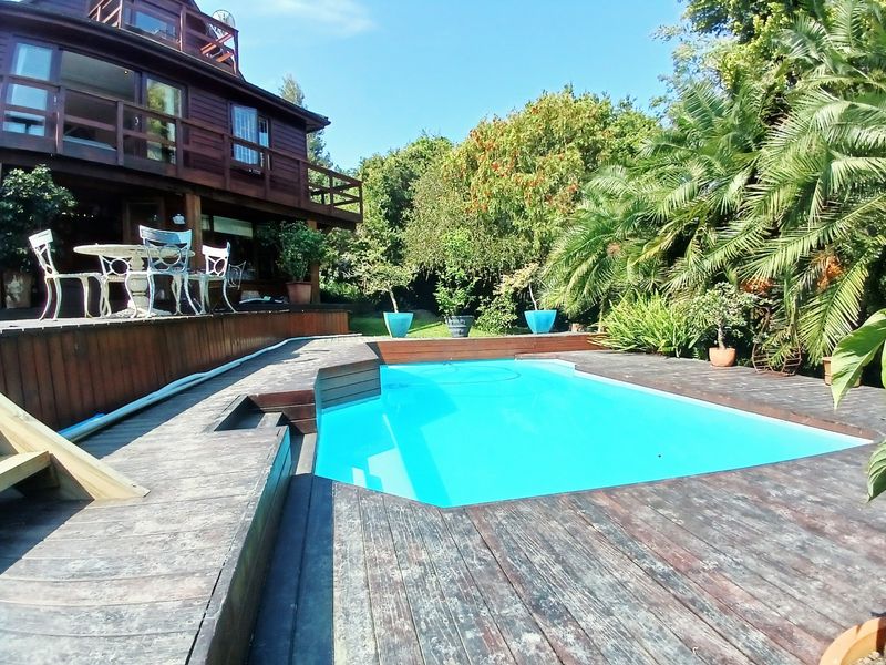 Outdoor pool area featuring extensive timber decking and a multi-level wooden house facade, set within a lush garden environment.