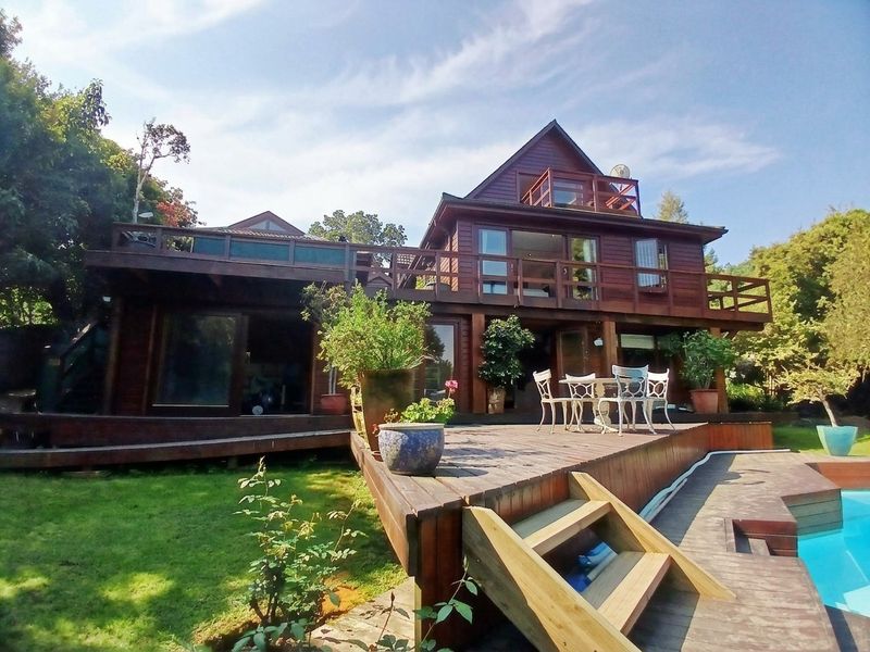 Rear view of a multi-level timber house featuring extensive wooden decking, balconies, and a swimming pool set within a garden.