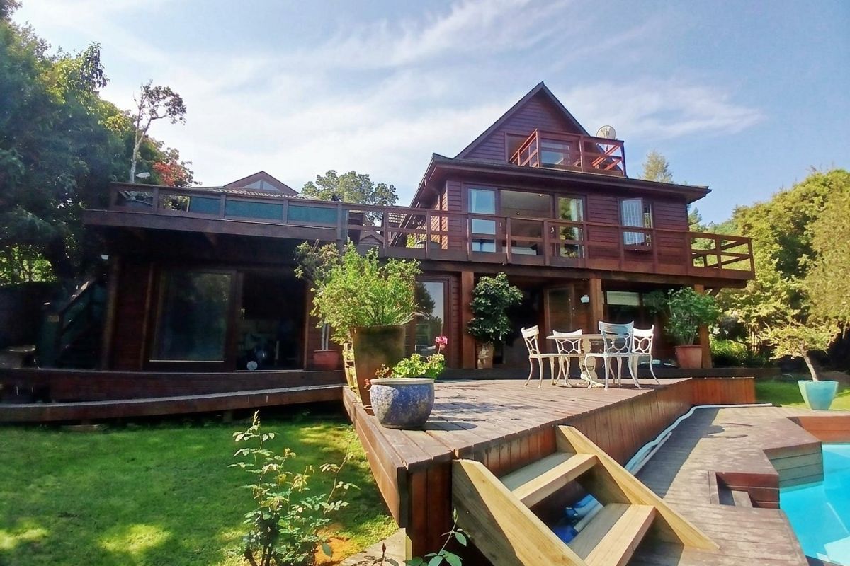 Rear view of a multi-level timber house featuring extensive wooden decking, balconies, and a swimming pool set within a garden.