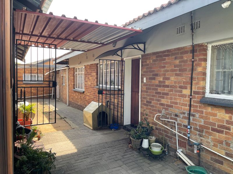 A rear exterior view of a brick house featuring a paved courtyard, security gates, and a covered walkway area.