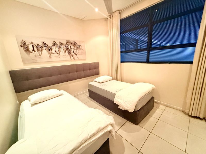 A well-lit bedroom featuring tiled flooring, a built-in upholstered headboard, and a large window with integrated blinds.