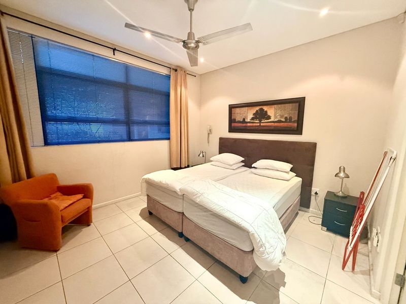 A functional bedroom featuring tiled flooring, a ceiling fan, and a large window with blinds, providing a neutral space ready for personalization.