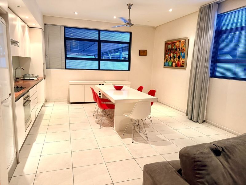 An open-plan kitchen and dining area featuring white cabinetry, tiled flooring, and modern built-in appliances, illuminated by recessed lighting and natural light from large windows.