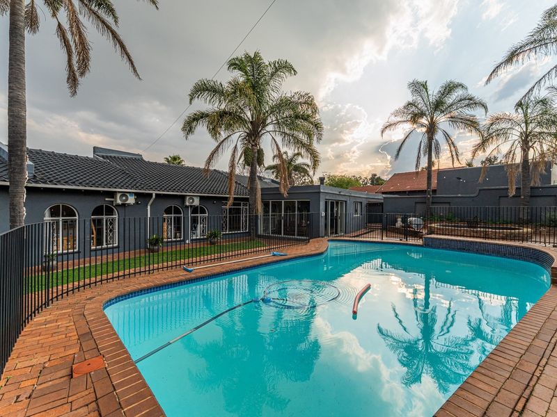 A well-maintained swimming pool area featuring brick coping, secure metal fencing, and a view of the rear building facade with arched windows.