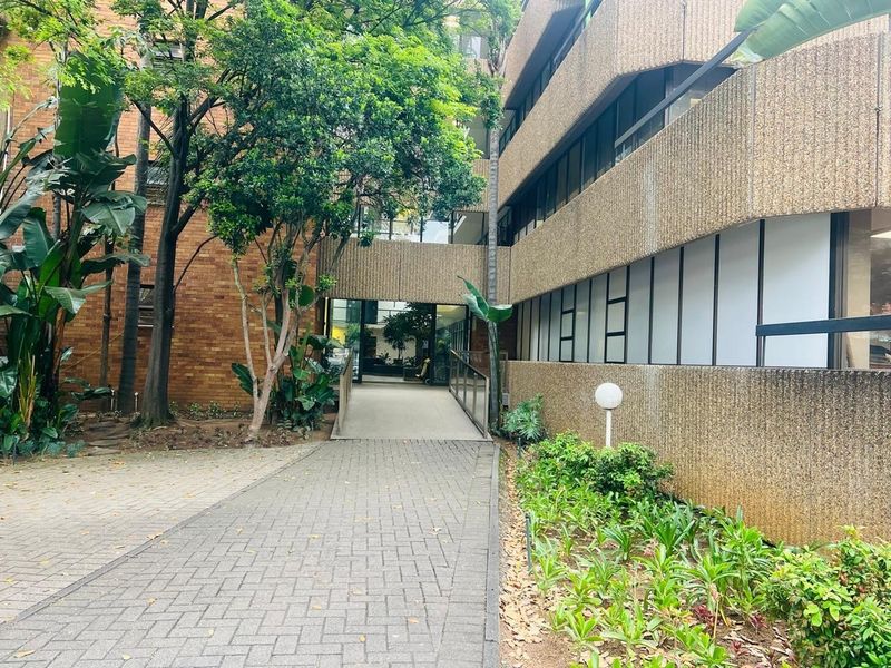 A paved walkway leading to the building entrance, featuring a ramp, landscaped garden areas, and mature trees surrounding the office complex exterior.