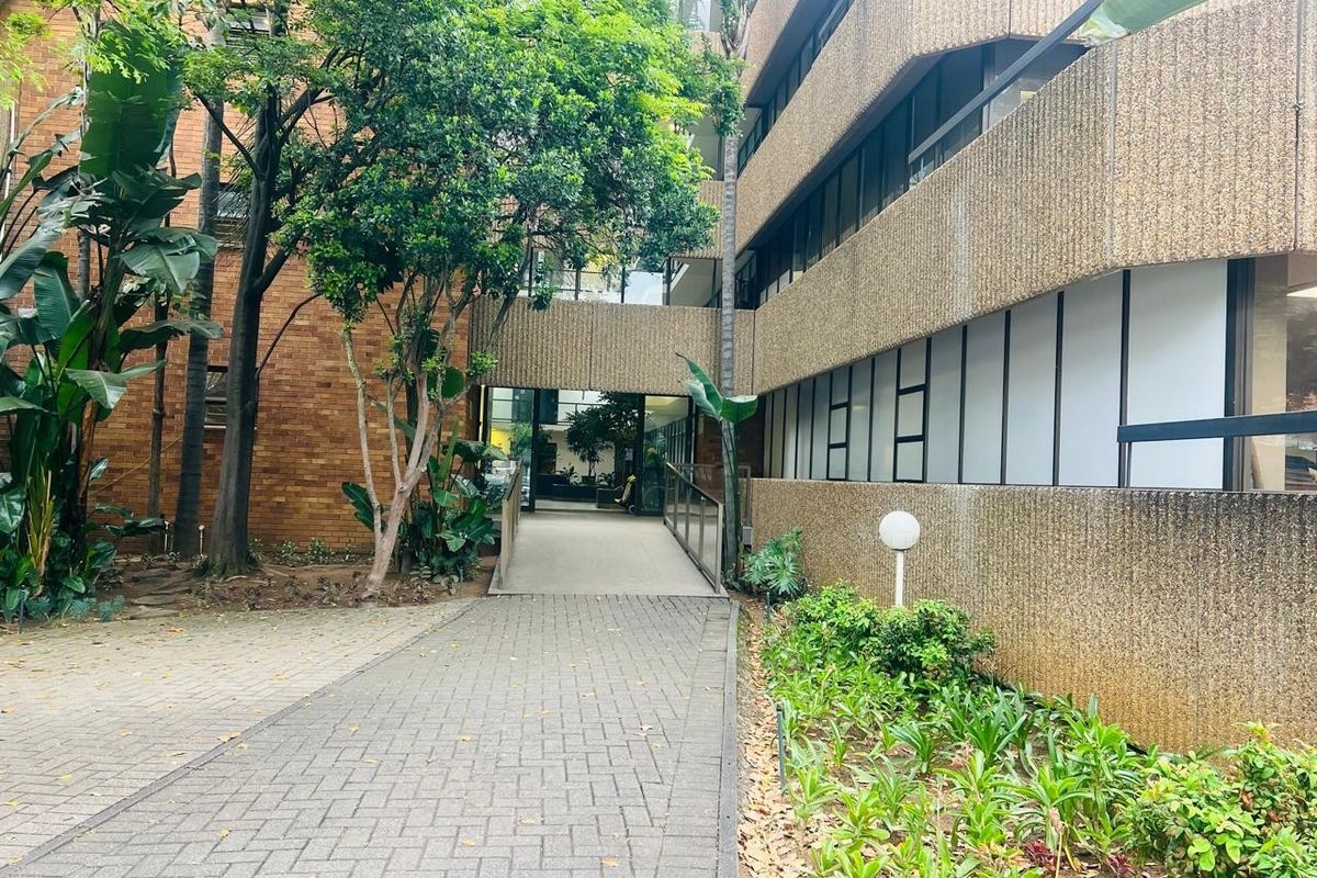 A paved walkway leading to the building entrance, featuring a ramp, landscaped garden areas, and mature trees surrounding the office complex exterior.