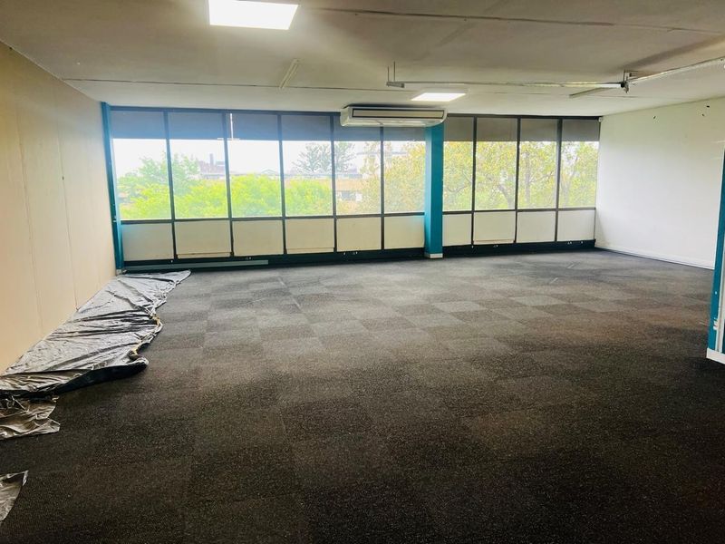 Spacious, empty office area featuring large windows, carpeted flooring, and ceiling-mounted air conditioning, offering a versatile workspace with natural light.