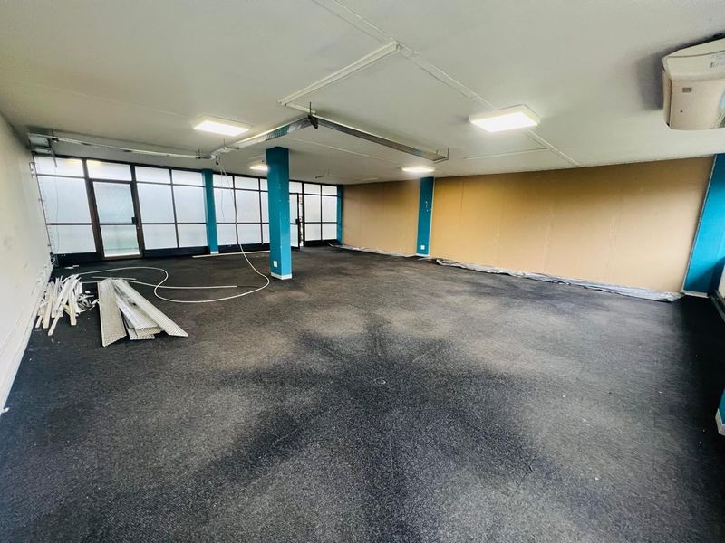 An open-plan office space featuring large windows, structural columns, and air conditioning, currently in need of refurbishment and flooring updates.