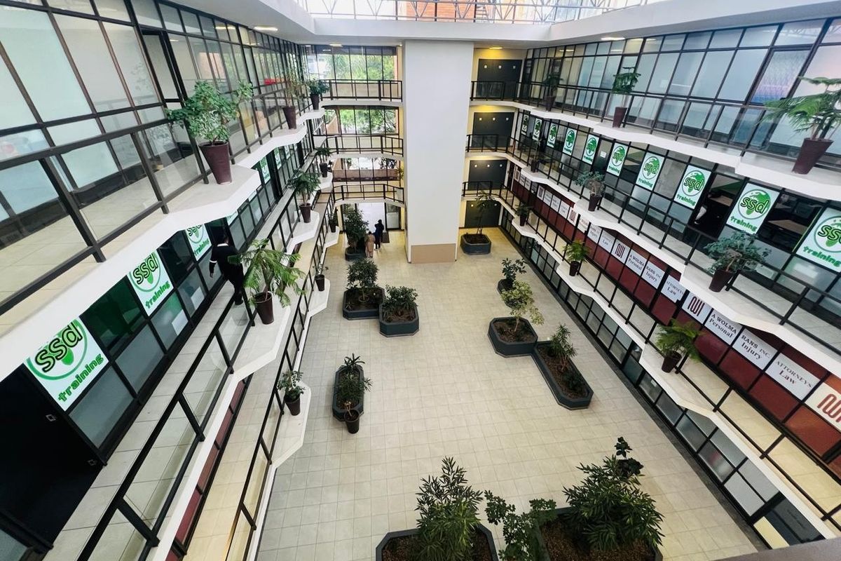A bright, multi-level office atrium featuring a large skylight, tiled flooring, and glass-walled office suites arranged around a central open space.