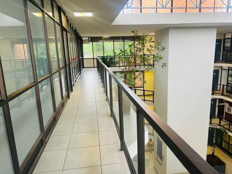 A bright, modern office hallway featuring tiled flooring, a glass balustrade overlooking the atrium, and extensive glass partitioning for natural light.