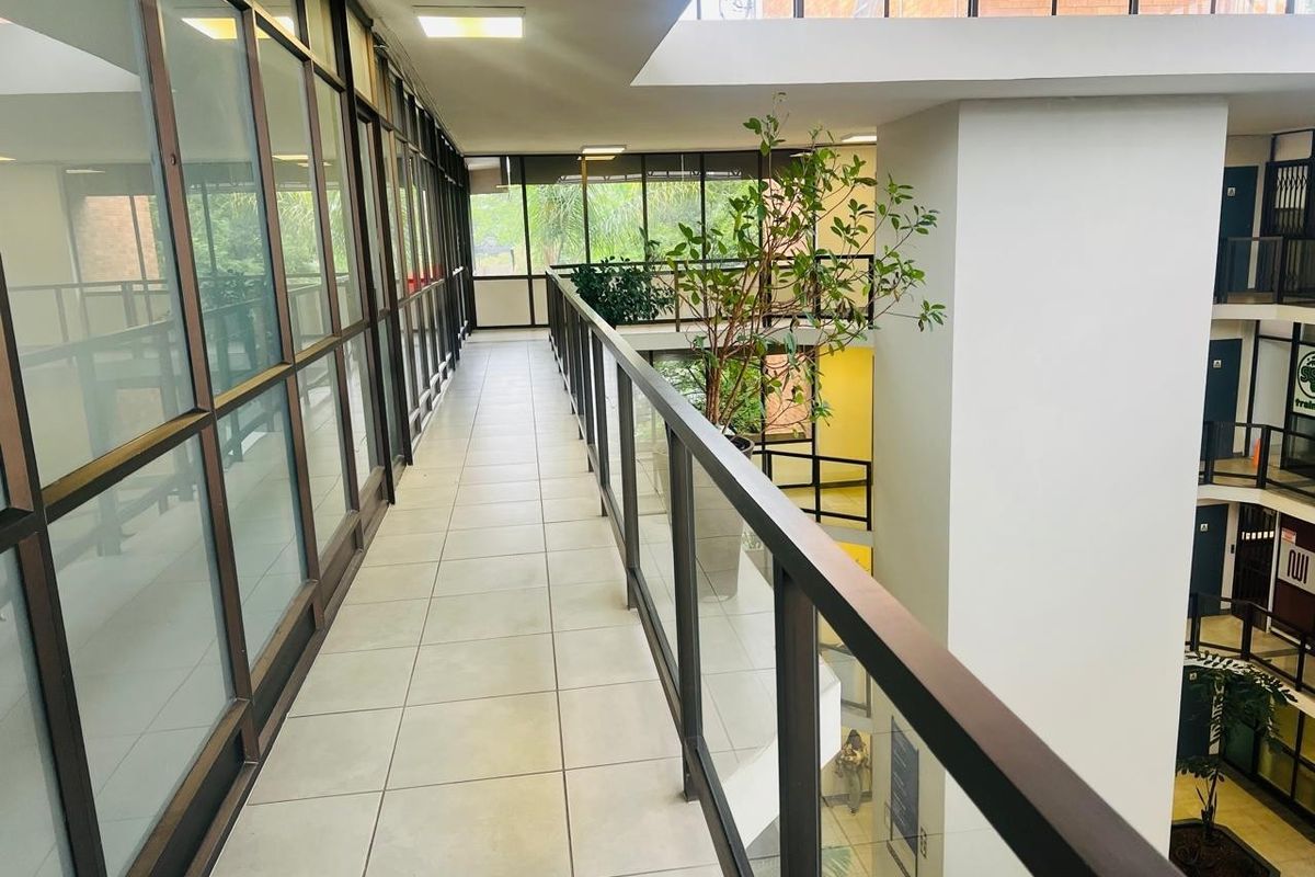 A bright, modern office hallway featuring tiled flooring, a glass balustrade overlooking the atrium, and extensive glass partitioning for natural light.