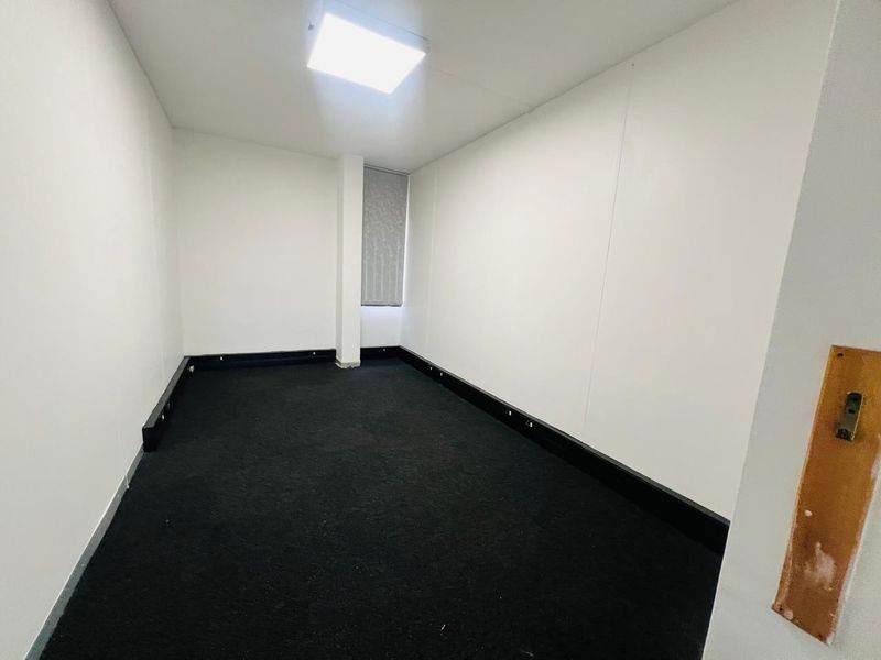 A vacant office space featuring dark carpeting, white walls, integrated skirting trunking for cabling, and a single window with a roller blind.