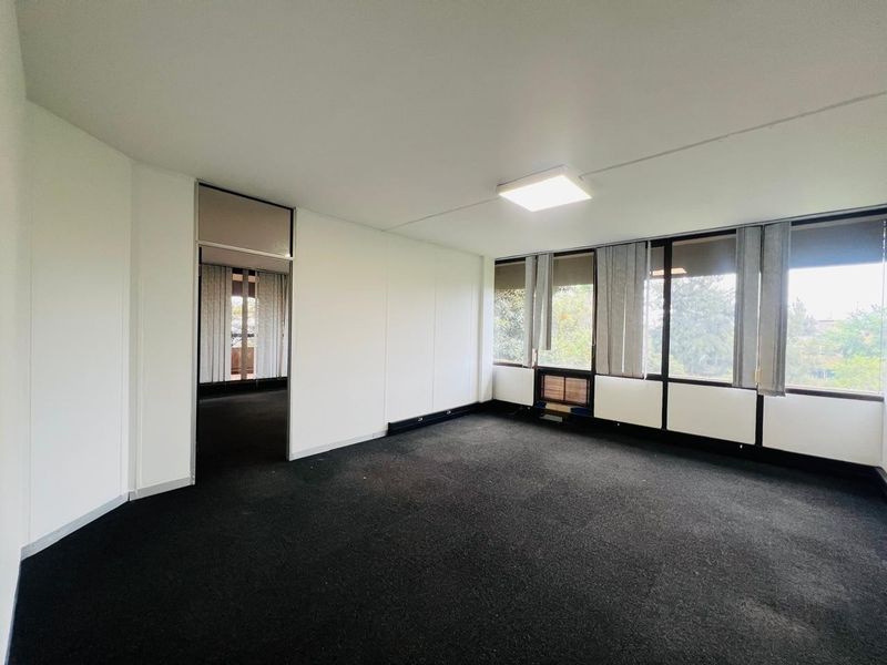 An empty, well-lit office space featuring black carpeting, large windows with vertical blinds, and a neutral interior finish.