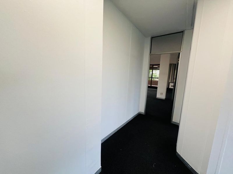 A clean, empty office hallway featuring dark carpeting and white partition walls, leading into an adjacent workspace.