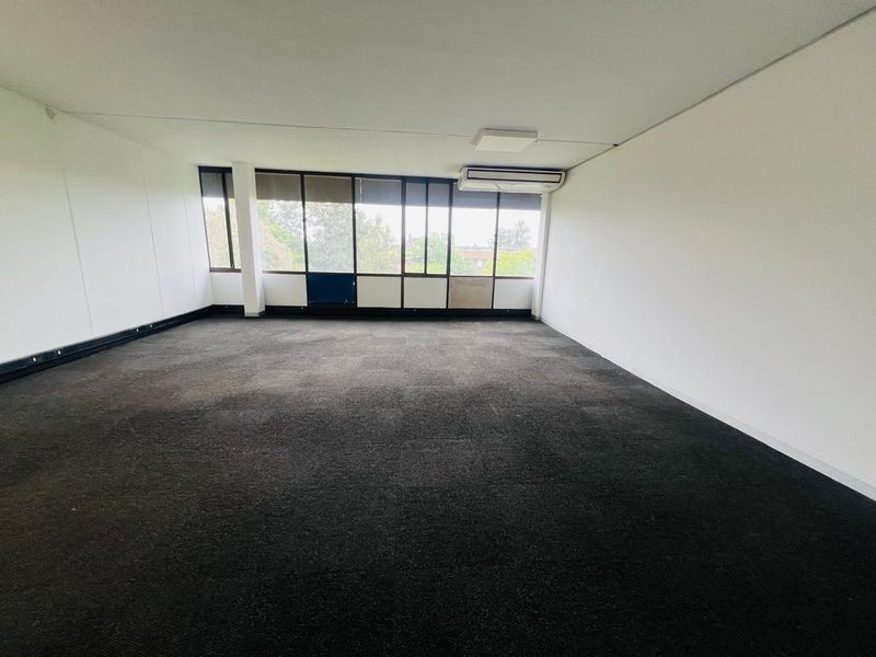 A spacious, empty office area featuring large windows, wall-mounted air conditioning, and dark carpeting, offering a blank canvas for professional workspace customization.