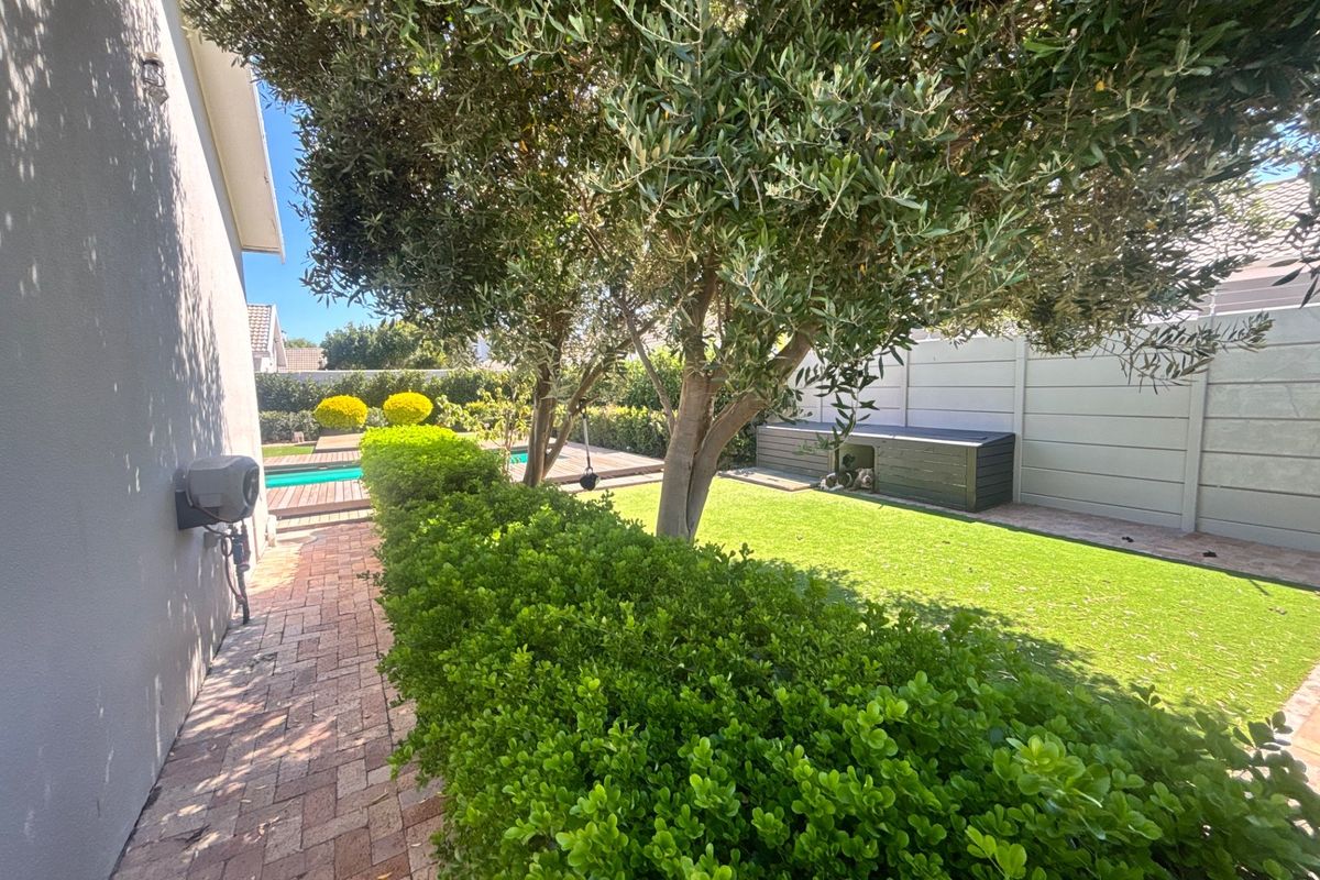 A well-maintained rear garden featuring a swimming pool, paved pathways, mature trees, and a secure perimeter wall.