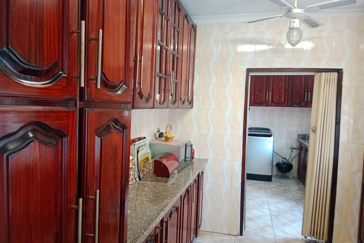 A functional kitchen featuring dark wood cabinetry, granite countertops, and tiled finishes, with an adjoining scullery area visible through the doorway.