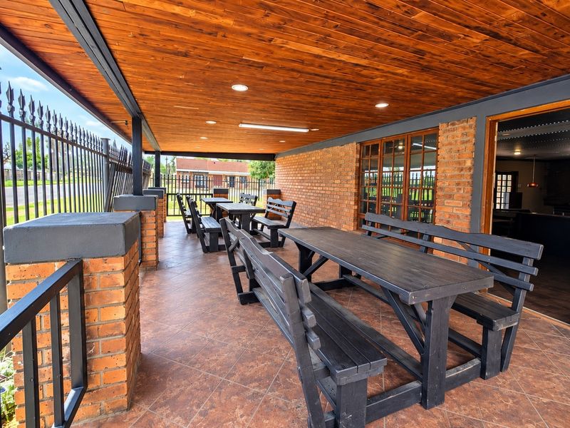 A spacious covered outdoor patio featuring a warm timber-clad ceiling, recessed lighting, and durable tiled flooring, enclosed by brick pillars and metal security fencing.
