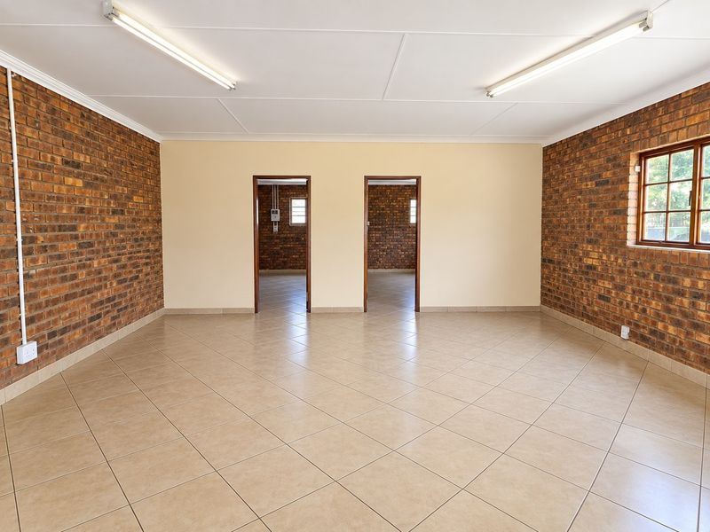 A spacious, empty commercial office area featuring exposed brick walls, neutral ceramic floor tiles, and bright overhead fluorescent lighting.
