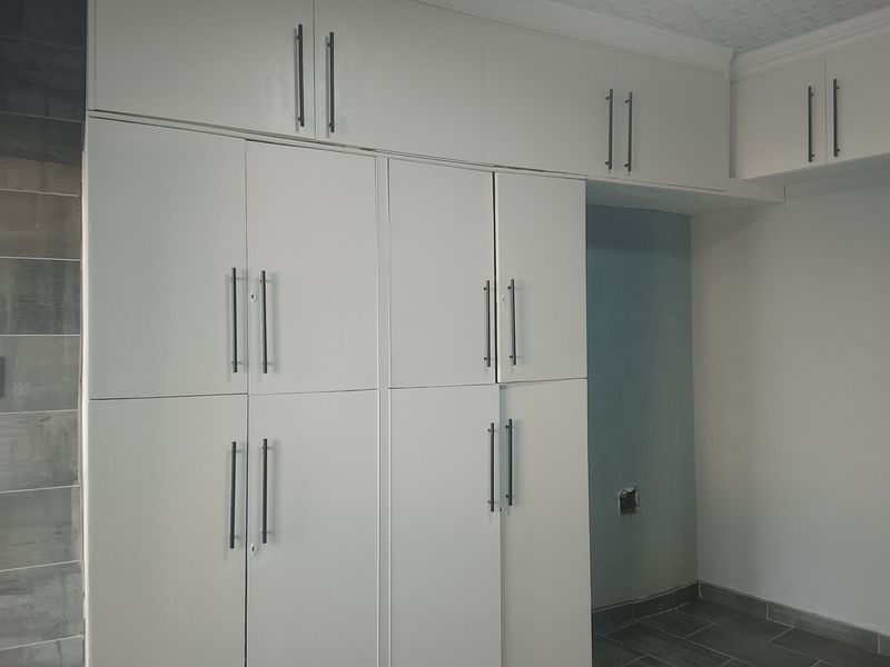 A kitchen area featuring white built-in cabinetry with modern handles and tiled flooring, currently vacant and awaiting final finishes.