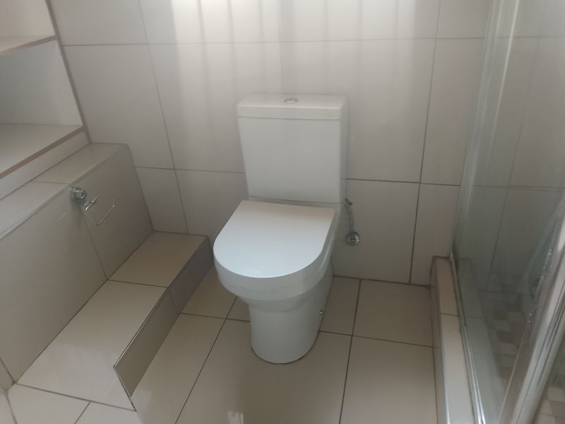 A clean, functional bathroom featuring a modern toilet, tiled finishes, and a glass-enclosed shower area with built-in storage shelving.