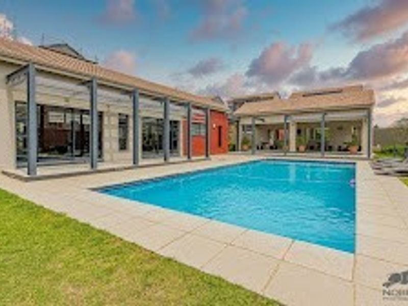 A well-maintained outdoor pool area featuring a clear swimming pool, tiled decking, and a covered patio structure attached to the main residence.