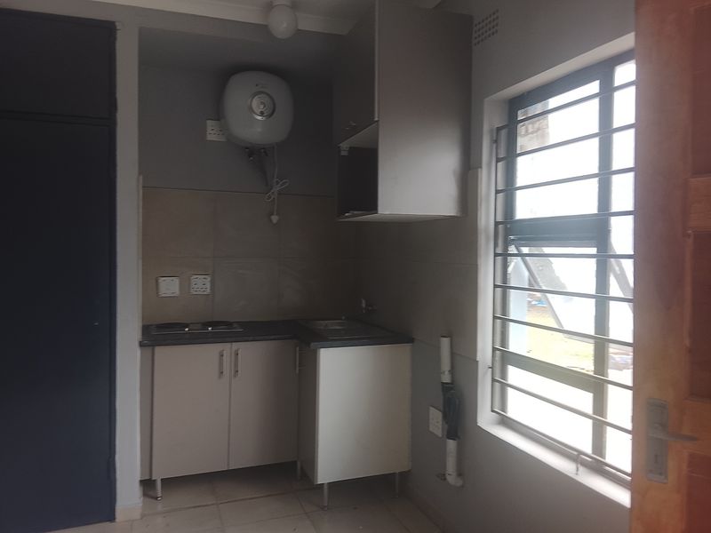 Compact kitchen area featuring built-in cabinetry, a sink, an electric hob, and an electric water heater, with tiled flooring and security bars on the window.