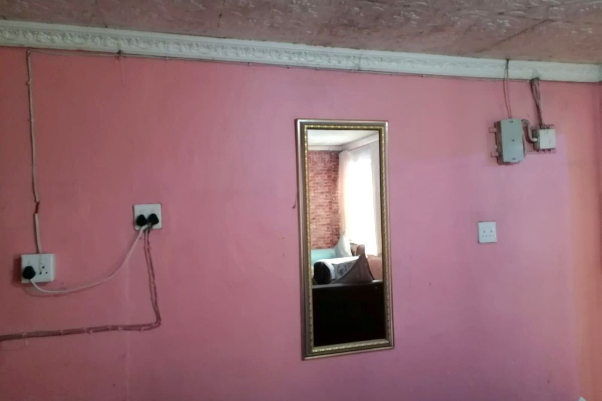 A bedroom interior featuring pink walls, textured ceiling, and visible surface-mounted electrical wiring and outlets.