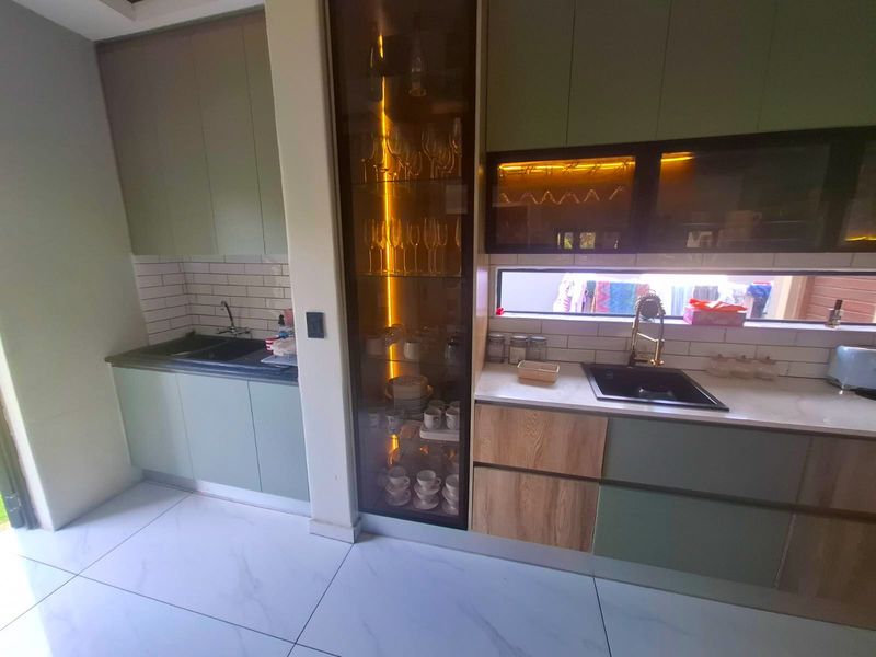 Modern kitchen featuring sage green and wood-tone cabinetry, subway tile backsplash, stone countertops, dual sinks, and integrated display shelving with accent lighting.