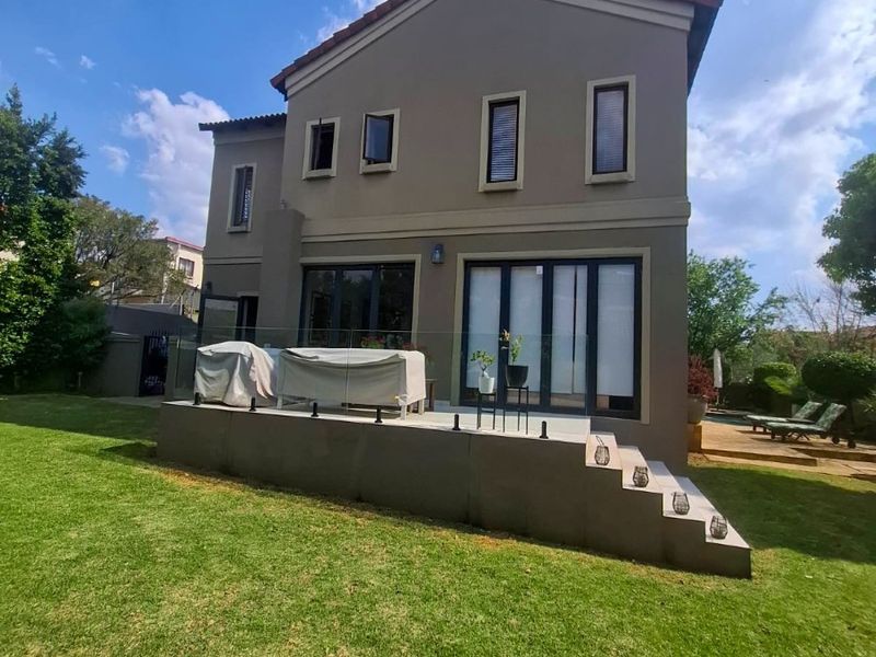 Rear view of a two-story house featuring a raised patio with glass balustrades, aluminium doors, and a well-maintained garden.