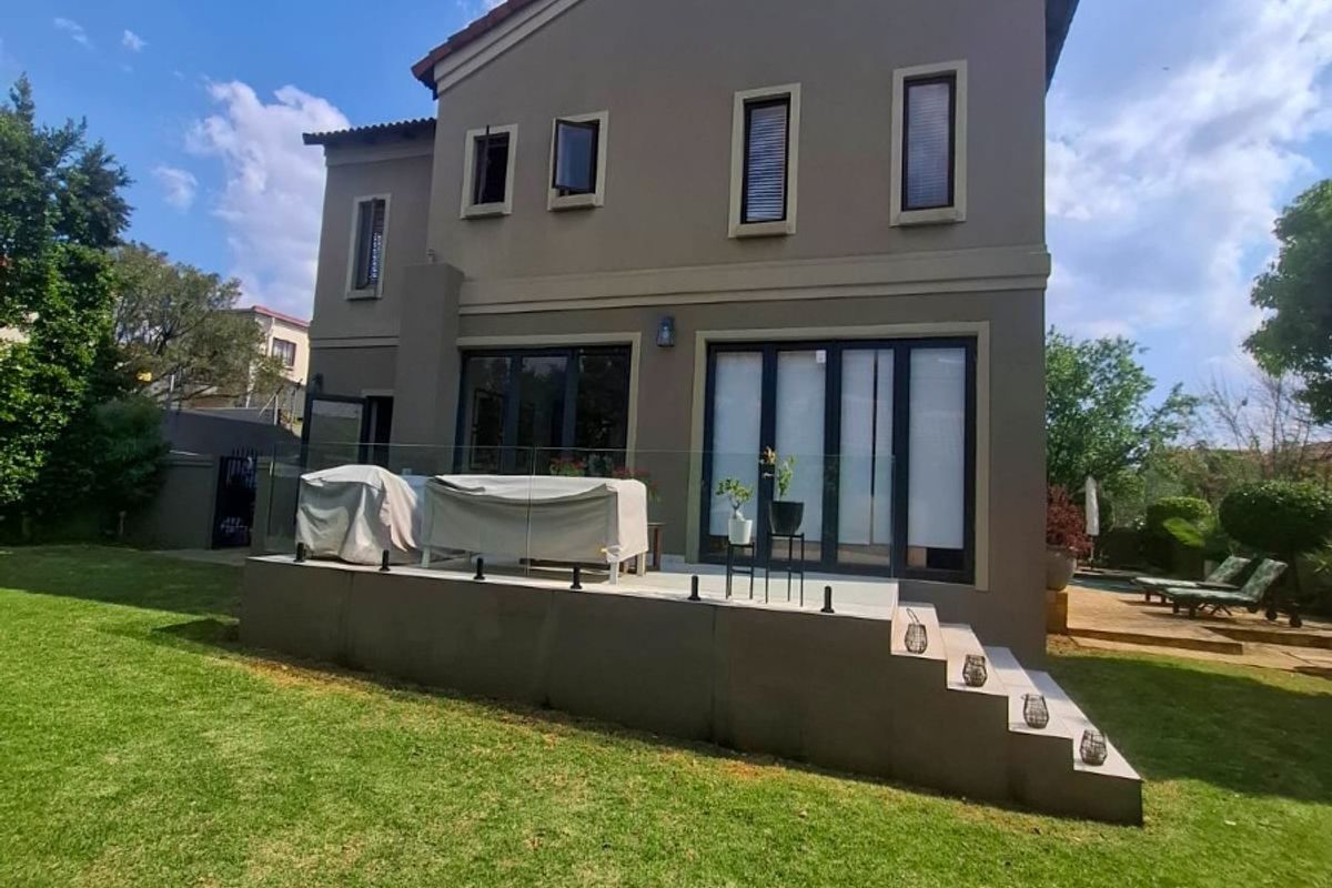 Rear view of a two-story house featuring a raised patio with glass balustrades, aluminium doors, and a well-maintained garden.