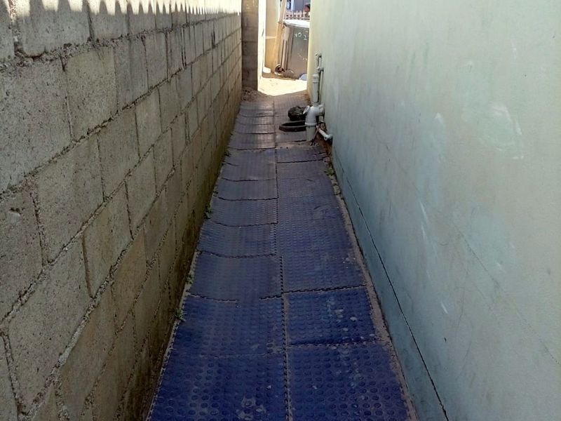 A narrow side passage between the house exterior and a concrete block boundary wall, featuring exposed plumbing and basic ground surfacing.
