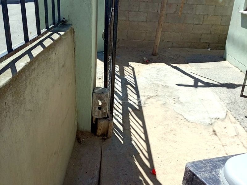 A narrow exterior entrance area featuring a metal security gate, perimeter walling, and concrete paving in need of maintenance.