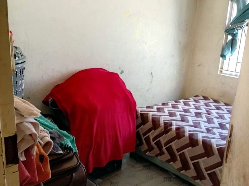 A compact bedroom featuring tiled flooring, a window with security bars, and wall ventilation, currently showing signs of wear and tear.