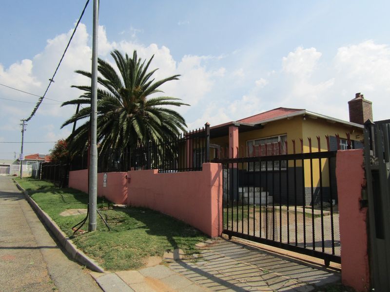 Front exterior view of a single-story house featuring a metal security gate, perimeter wall, and a prominent palm tree in the front yard.