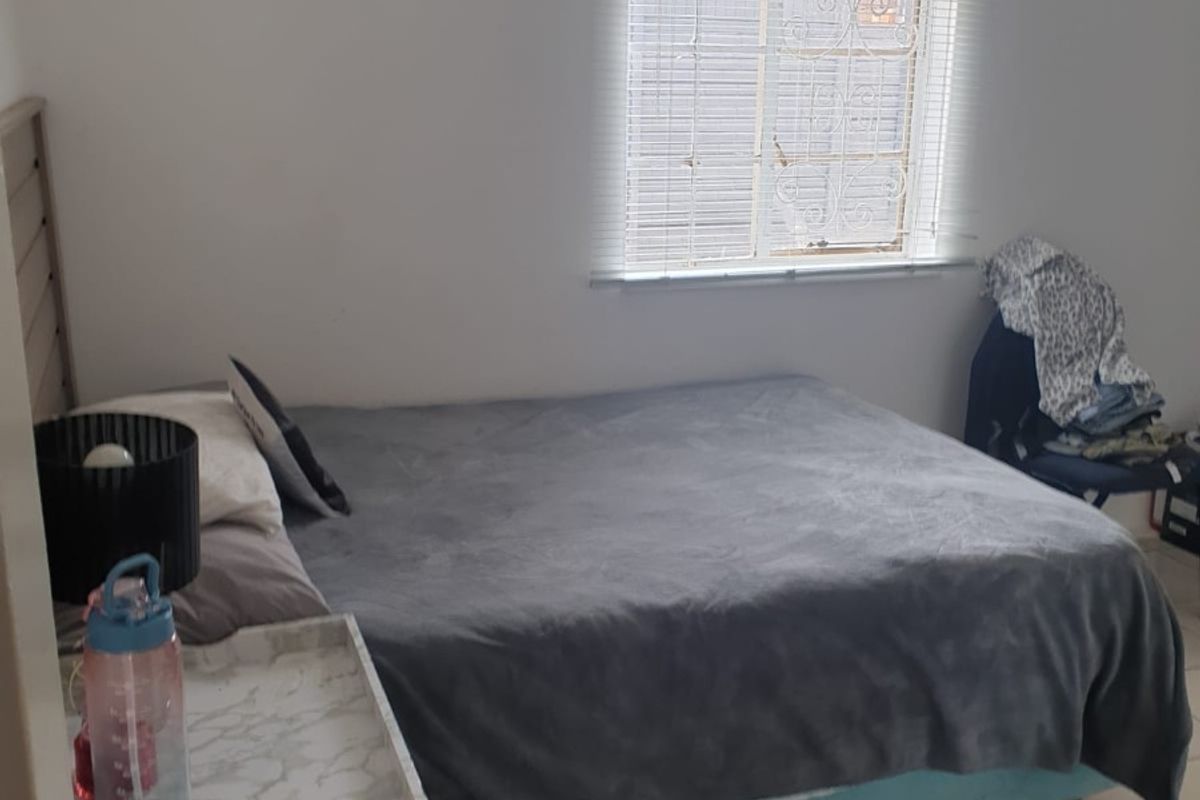 A functional bedroom featuring tiled flooring, a window with security bars, and venetian blinds, offering a simple and practical living space.