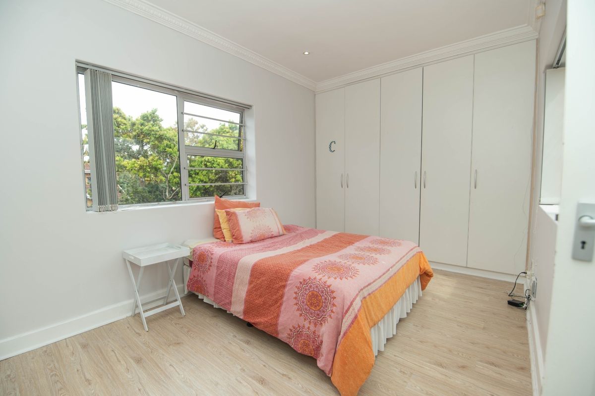 A bright bedroom featuring light-toned laminate flooring, built-in cupboards, and a large window providing natural light and a view of the surrounding greenery.