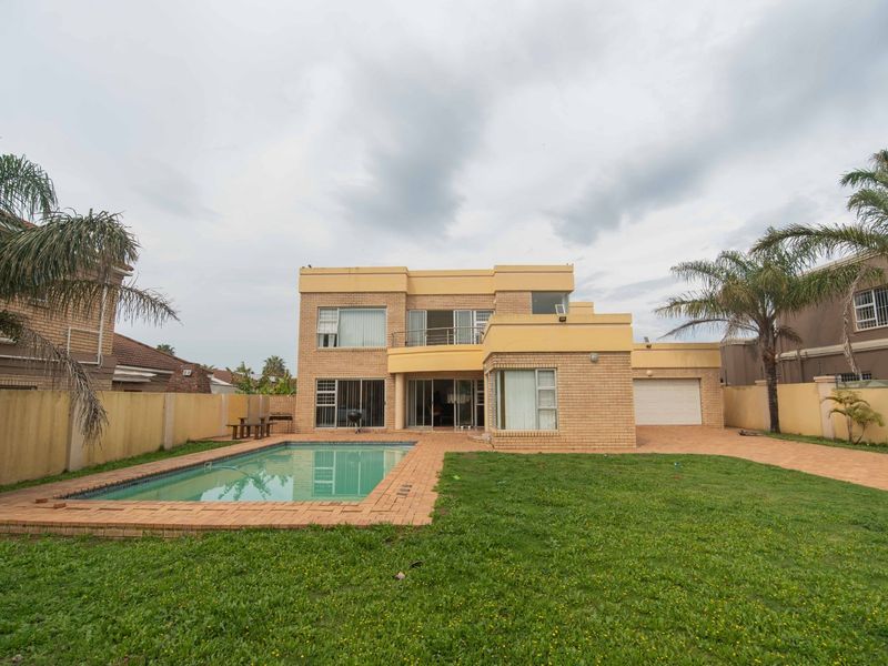 Rear view of a double-story brick house featuring a swimming pool, paved patio area, balcony, and a private garden with boundary walls.