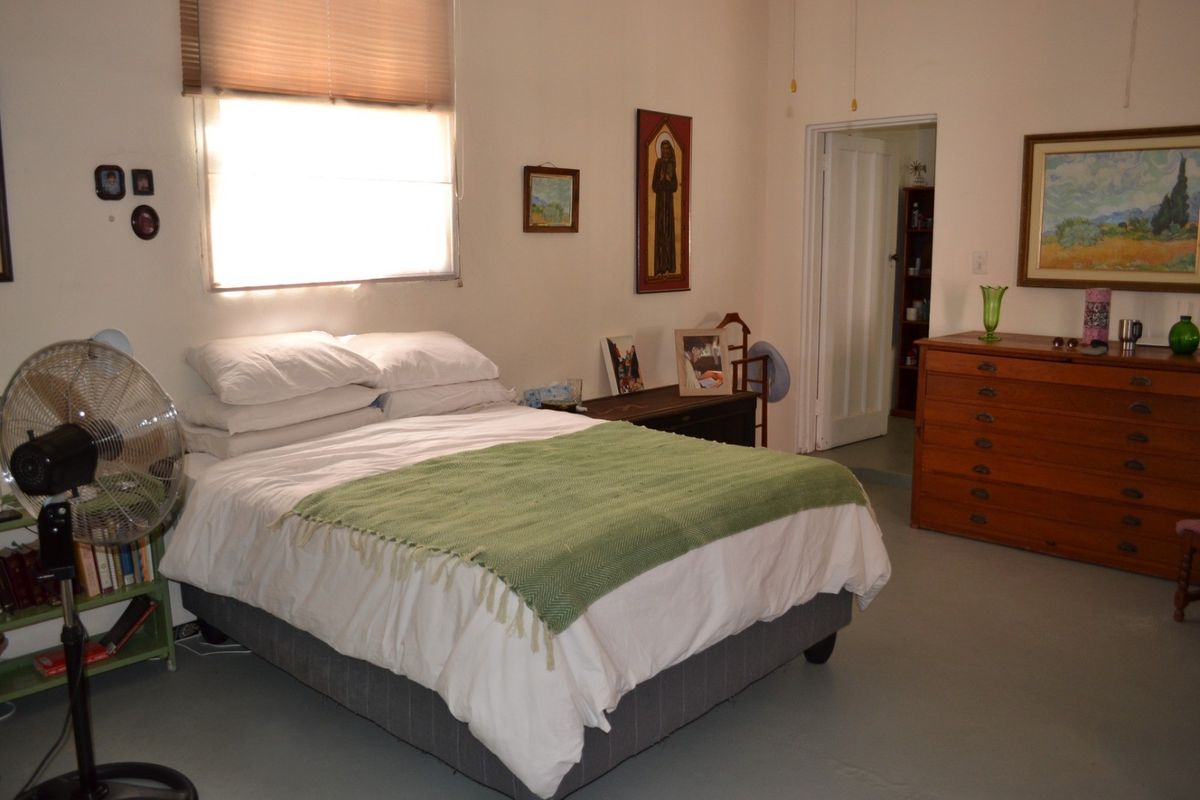 A functional bedroom featuring a window with a roller blind, painted concrete flooring, and access to an adjoining room via a standard internal door.