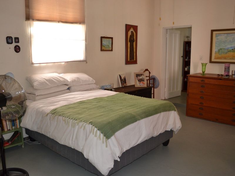 A functional bedroom featuring a window with a roller blind, painted concrete flooring, and access to an adjoining room via a standard internal door.