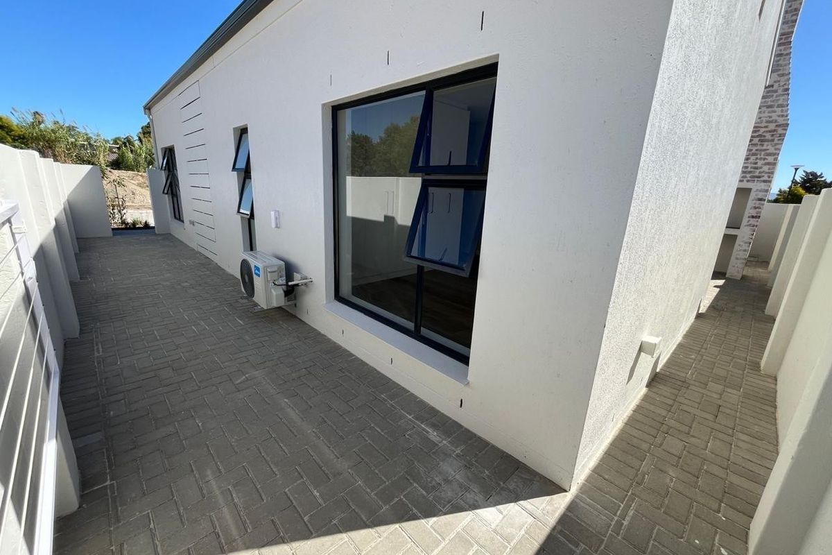 A clean, modern rear exterior view featuring paved pathways, rendered walls, and aluminium-framed windows with an external air conditioning unit.