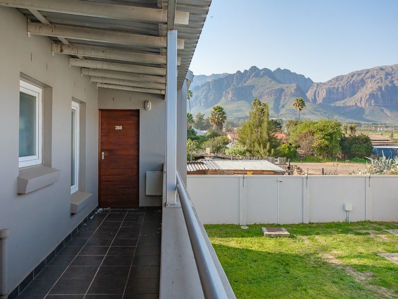 A covered balcony walkway featuring tiled flooring and metal railings, offering scenic mountain views and access to the apartment entrance.