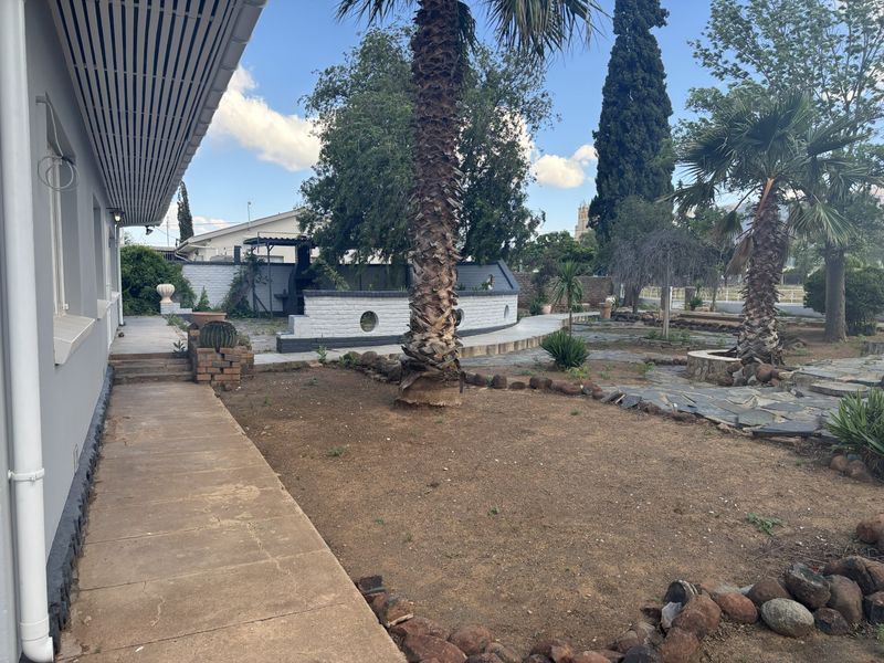 Spacious rear garden featuring a concrete patio, built-in braai area, and stone-paved pathways, offering potential for landscaping and outdoor entertainment development.