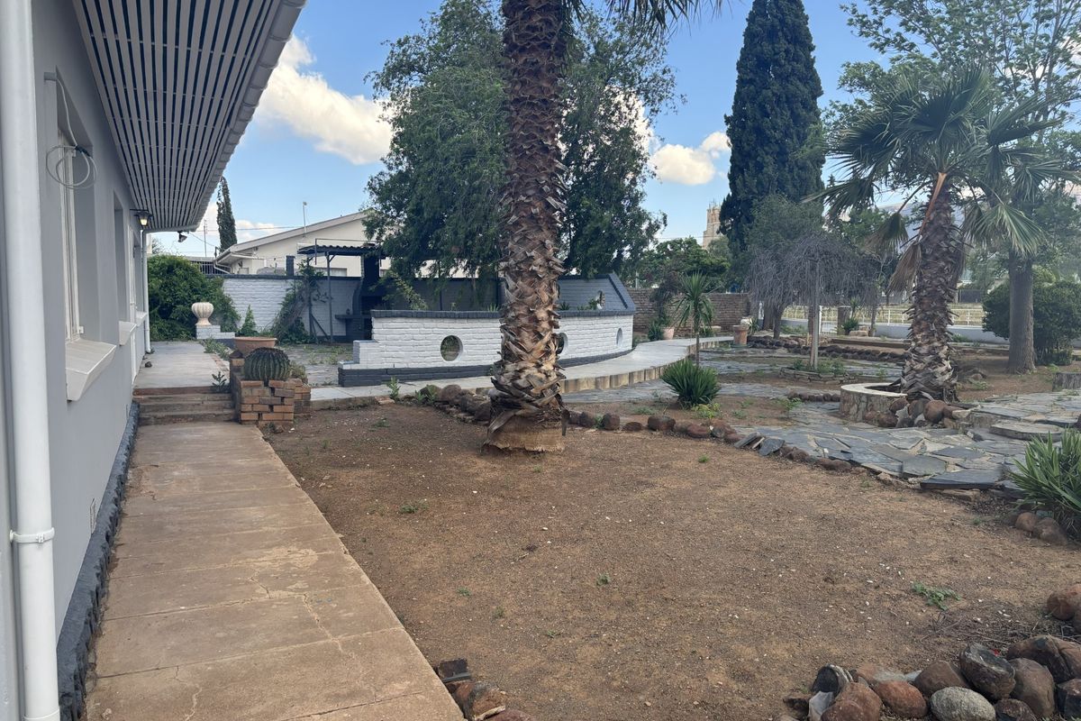 Spacious rear garden featuring a concrete patio, built-in braai area, and stone-paved pathways, offering potential for landscaping and outdoor entertainment development.