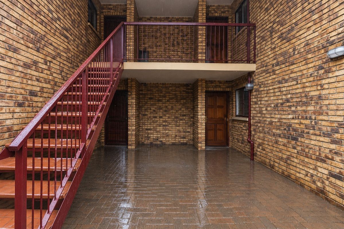 Exterior view of an apartment complex featuring a brick facade, paved courtyard area, and a metal staircase leading to an upper level balcony.