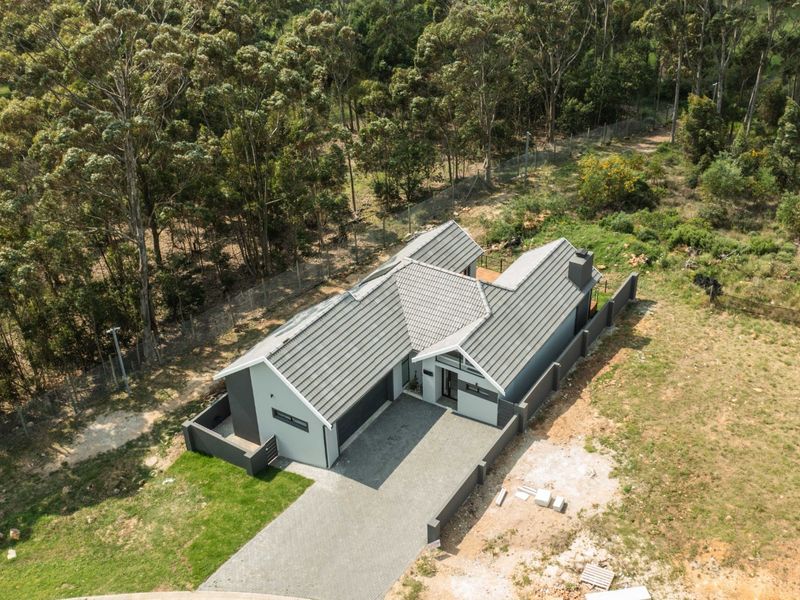 Aerial view of a modern house with a tiled roof, paved driveway, and surrounding boundary walls, set against a wooded landscape.