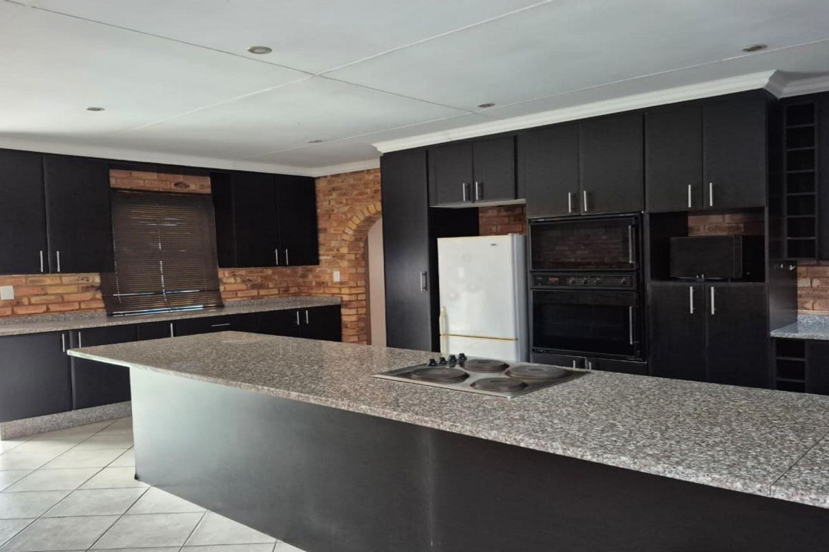 Spacious kitchen featuring dark cabinetry, granite countertops, a central island with a built-in hob, and exposed brick accents.