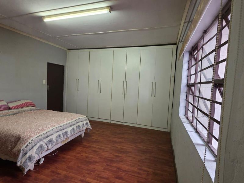 A bedroom featuring extensive built-in cupboards, laminate flooring, and security bars on the window. The room requires minor cosmetic updates to the ceiling and walls.
