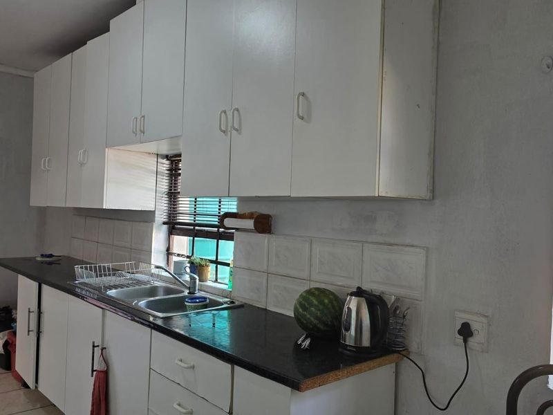 A functional kitchen featuring white built-in cabinetry, dark laminate countertops, a stainless steel double sink, and tiled backsplash with natural light from the window.