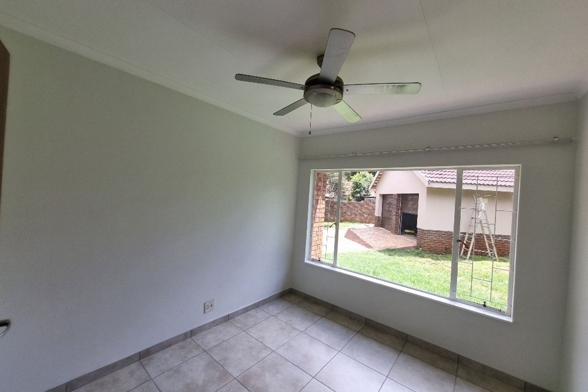 A bright, empty bedroom featuring neutral tiled flooring, a ceiling fan, and a large window overlooking the rear garden and garage.